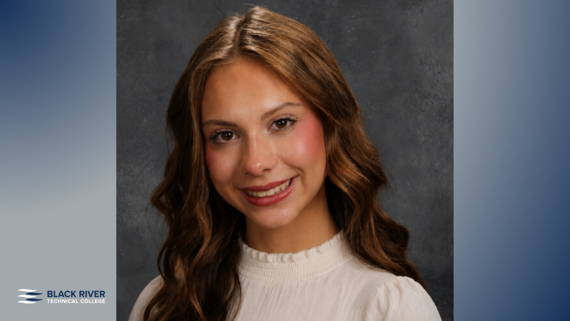 A young woman with long wavy brown hair smiles in a studio portrait, wearing a white ruffled-collar top against a gray backdrop.